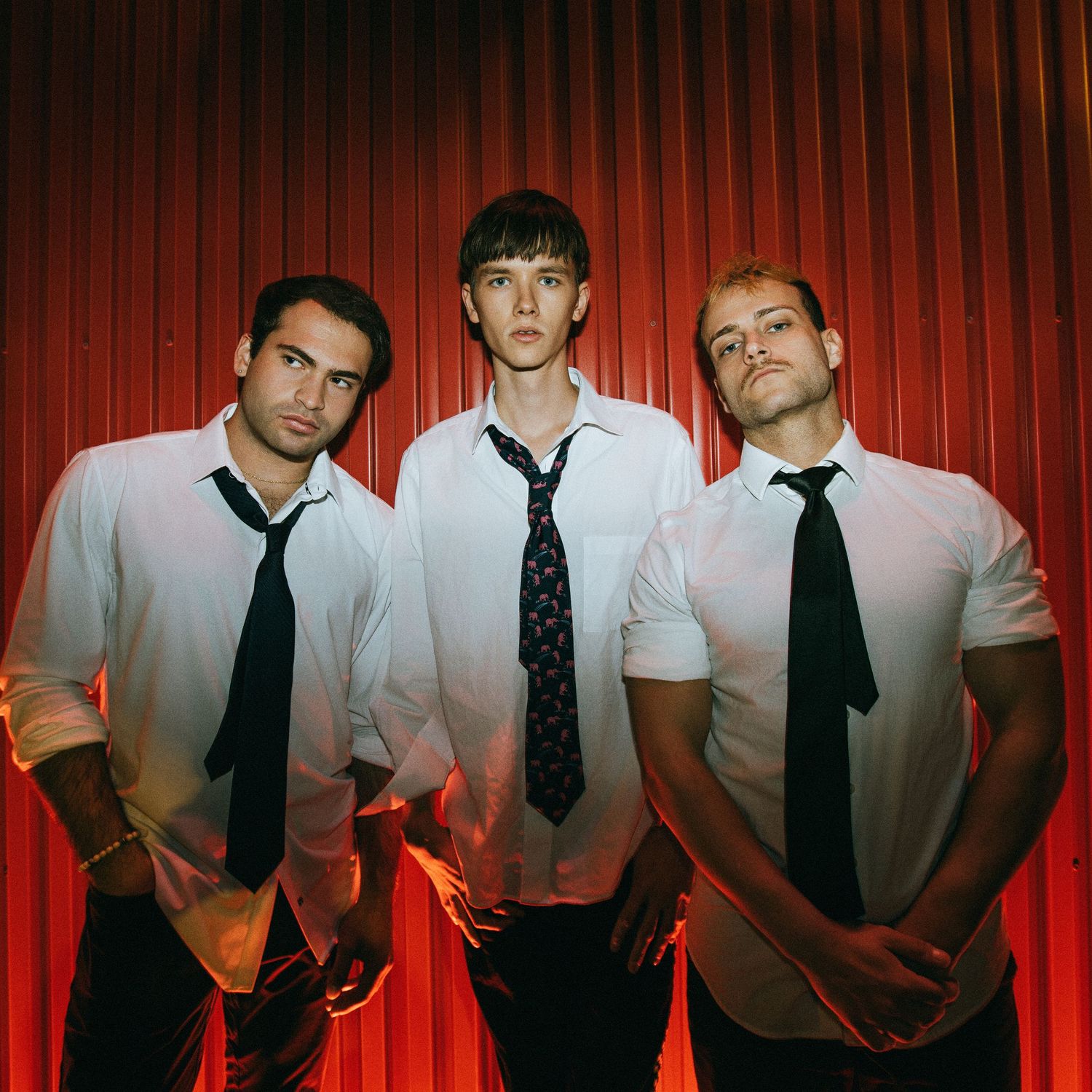 Three musicians in white dress shirts and black ties pose dramatically against a red curtain backdrop with moody lighting.