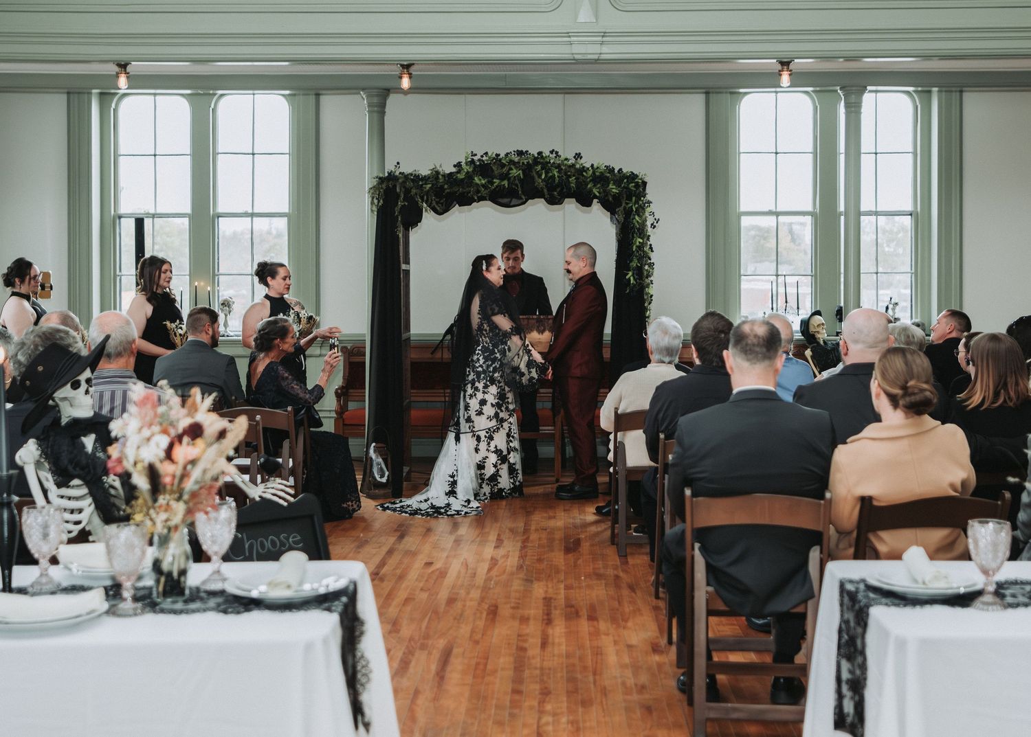 Wedding ceremony guests seated in rows at an elegant venue with arched windows and hardwood floors.