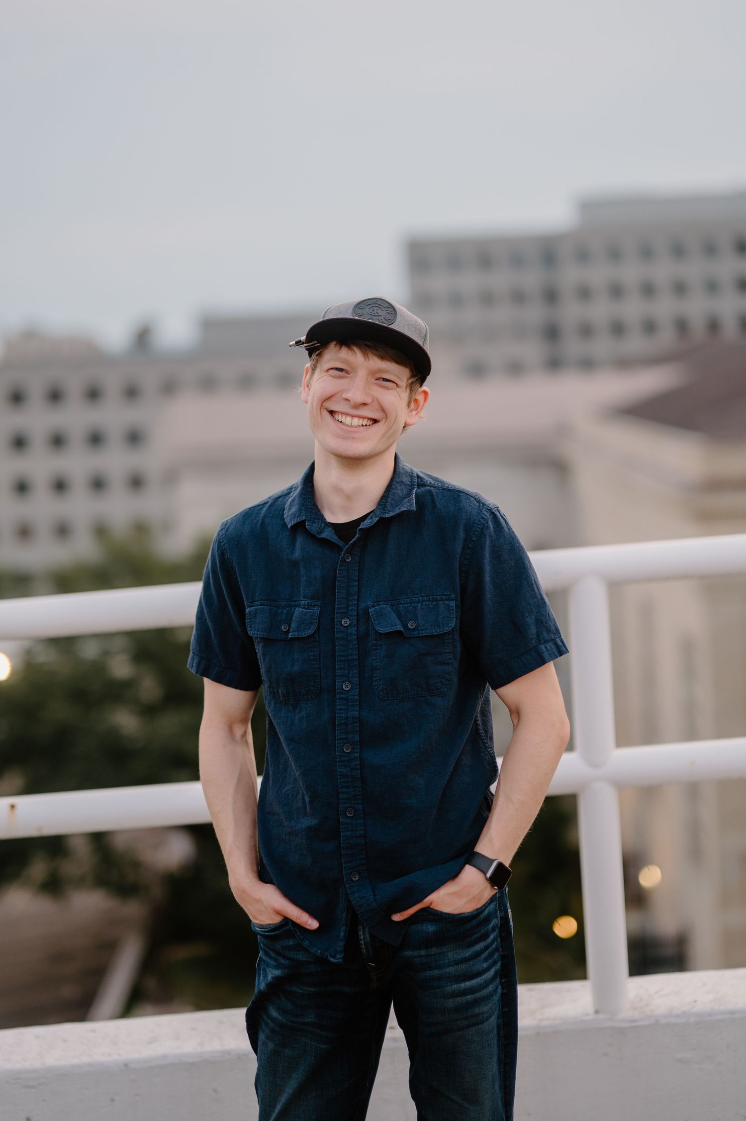 A person in a navy blue shirt and baseball cap poses on a balcony with urban buildings in the background.