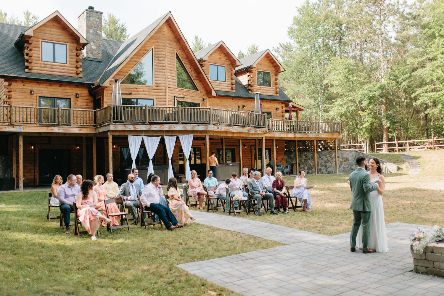 Outdoor wedding ceremony at rustic log cabin lodge with guests seated on lawn and couple dancing on stone patio.