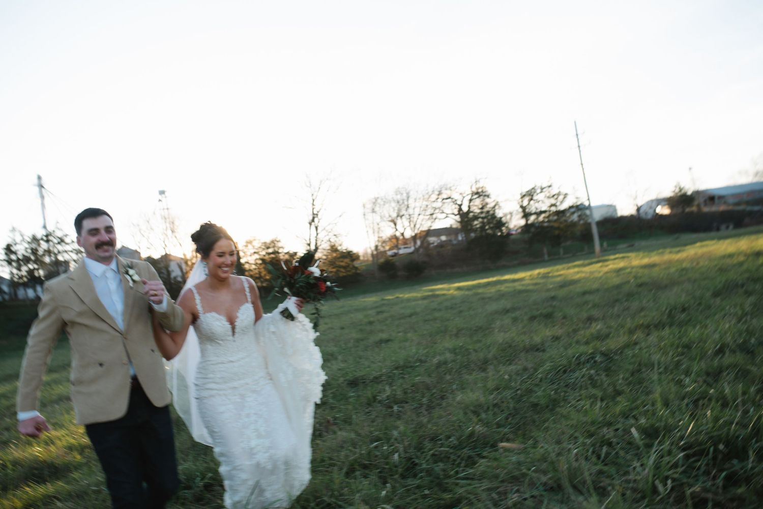 Couple walking together in formal attire across a grassy field at sunset.