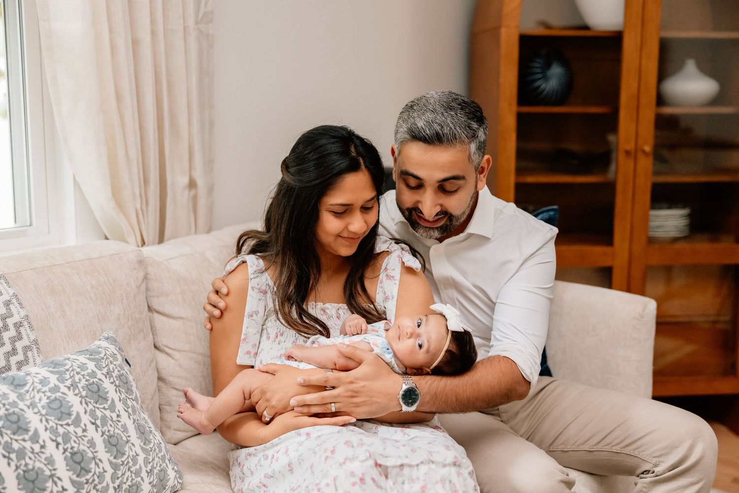 Two loving parents sitting on a beige couch tenderly hold and gaze at their newborn baby in a cozy living room.