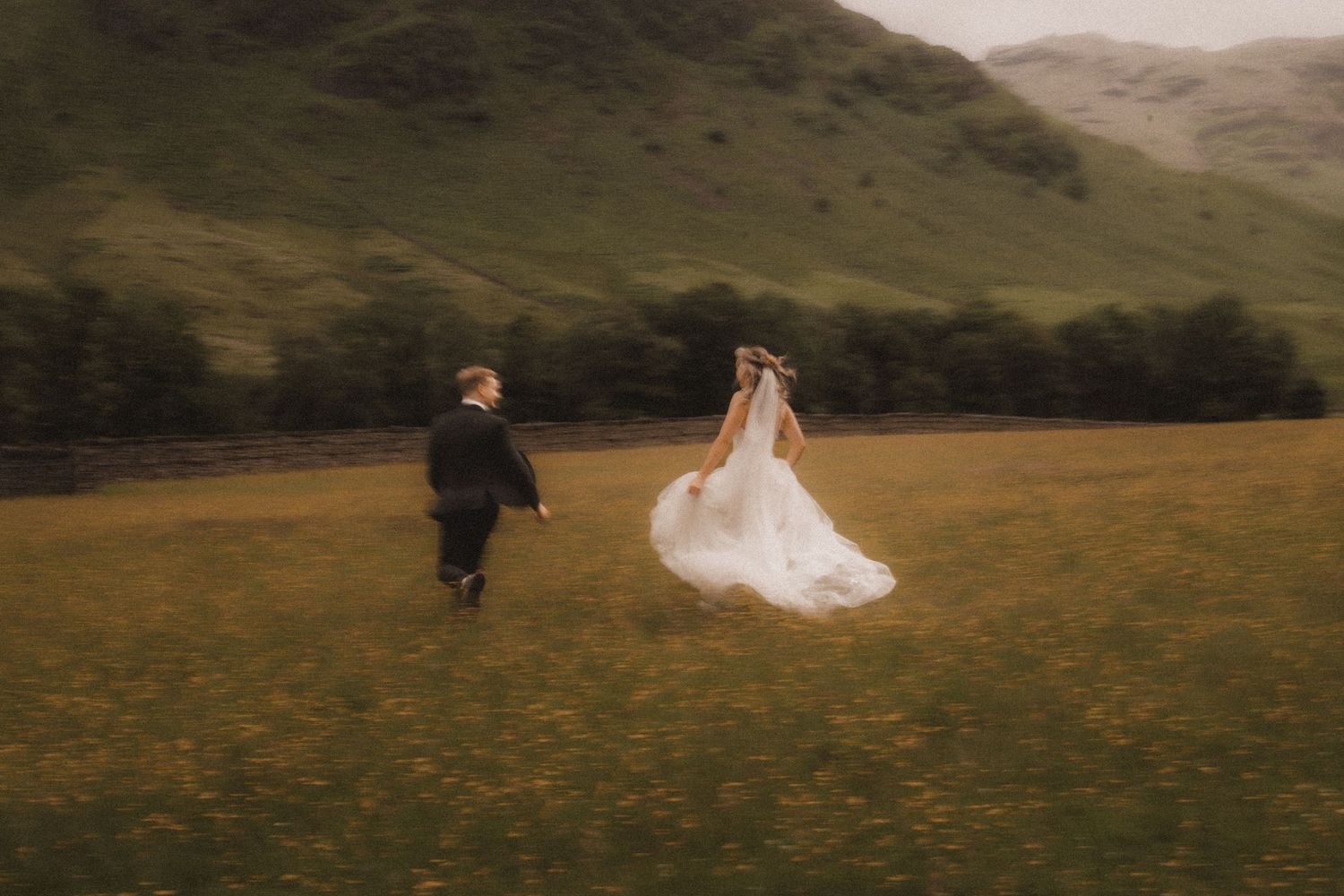 A couple runs through a misty field with rolling green hills in Lake District during their outdoor elopement wedding celebration.