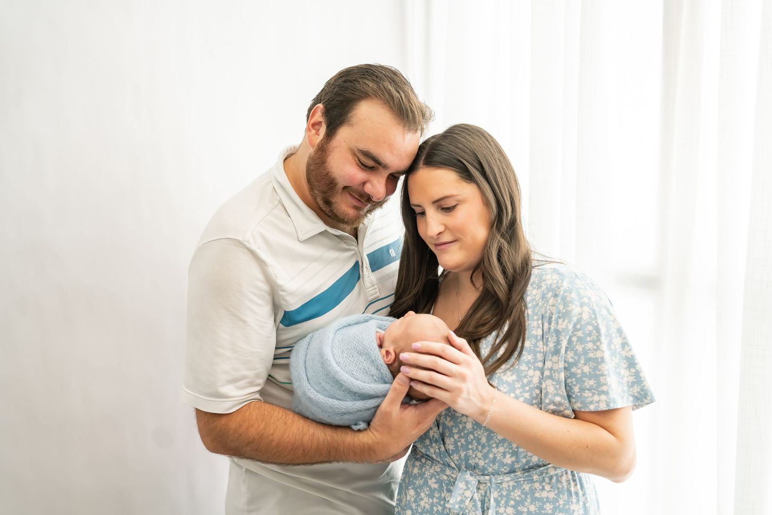 Two people in light clothing stand close together holding and looking lovingly at their newborn baby wrapped in blue.