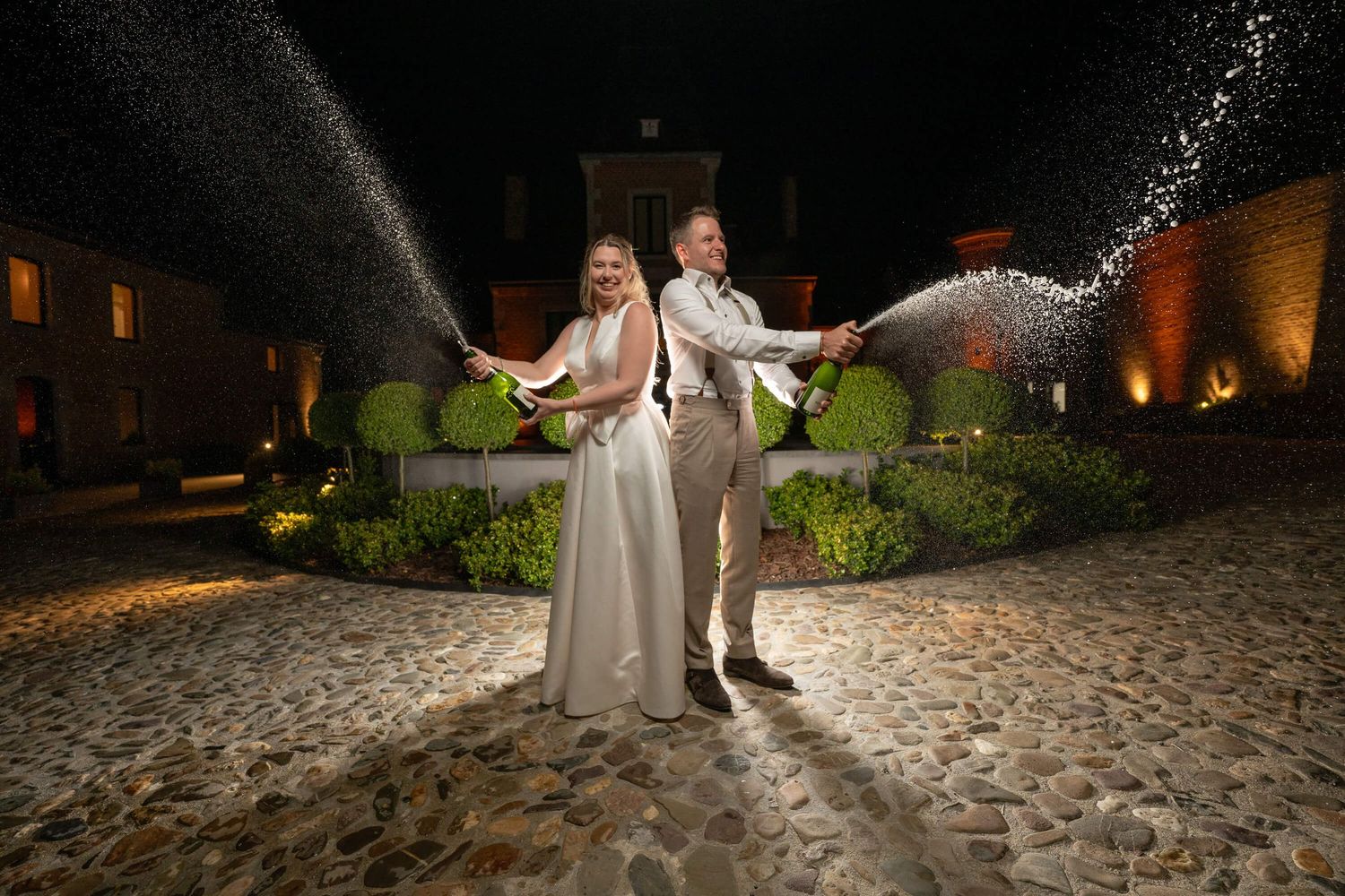 Two people in formal attire spray champagne at night in front of an illuminated stone building in a festive celebration.