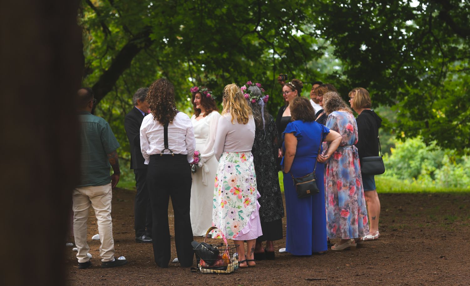 A group of people stand together in conversation on a dirt path surrounded by lush green trees.