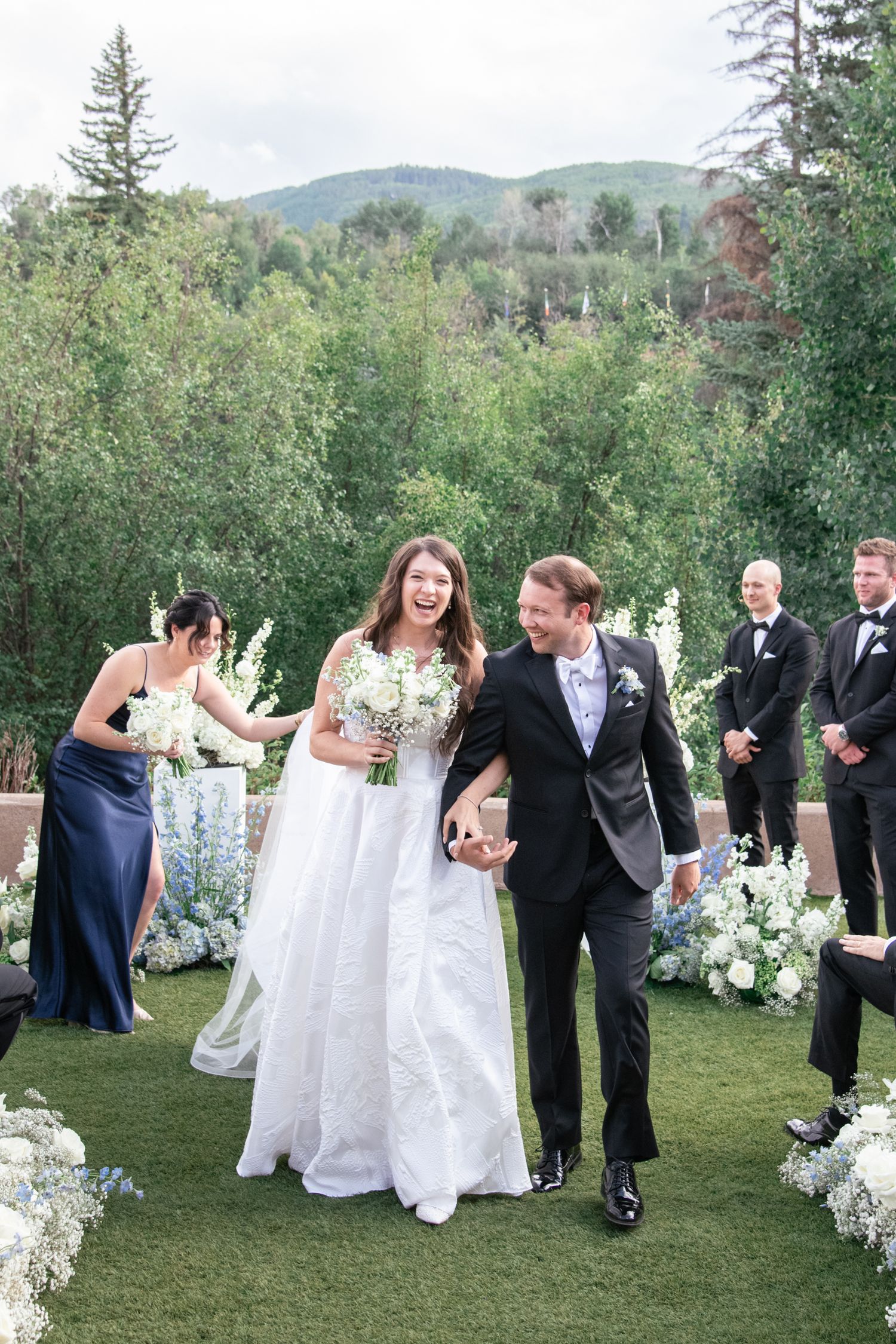 Wedding party laughing and celebrating on a manicured lawn with white floral arrangements and mountain backdrop.