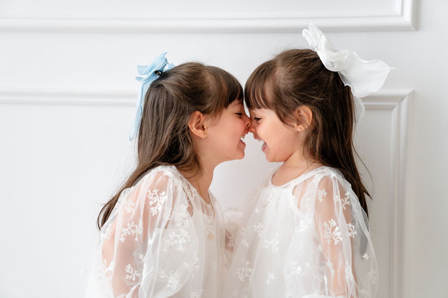 Two children in matching white lace dresses touch noses while smiling against a white wall.