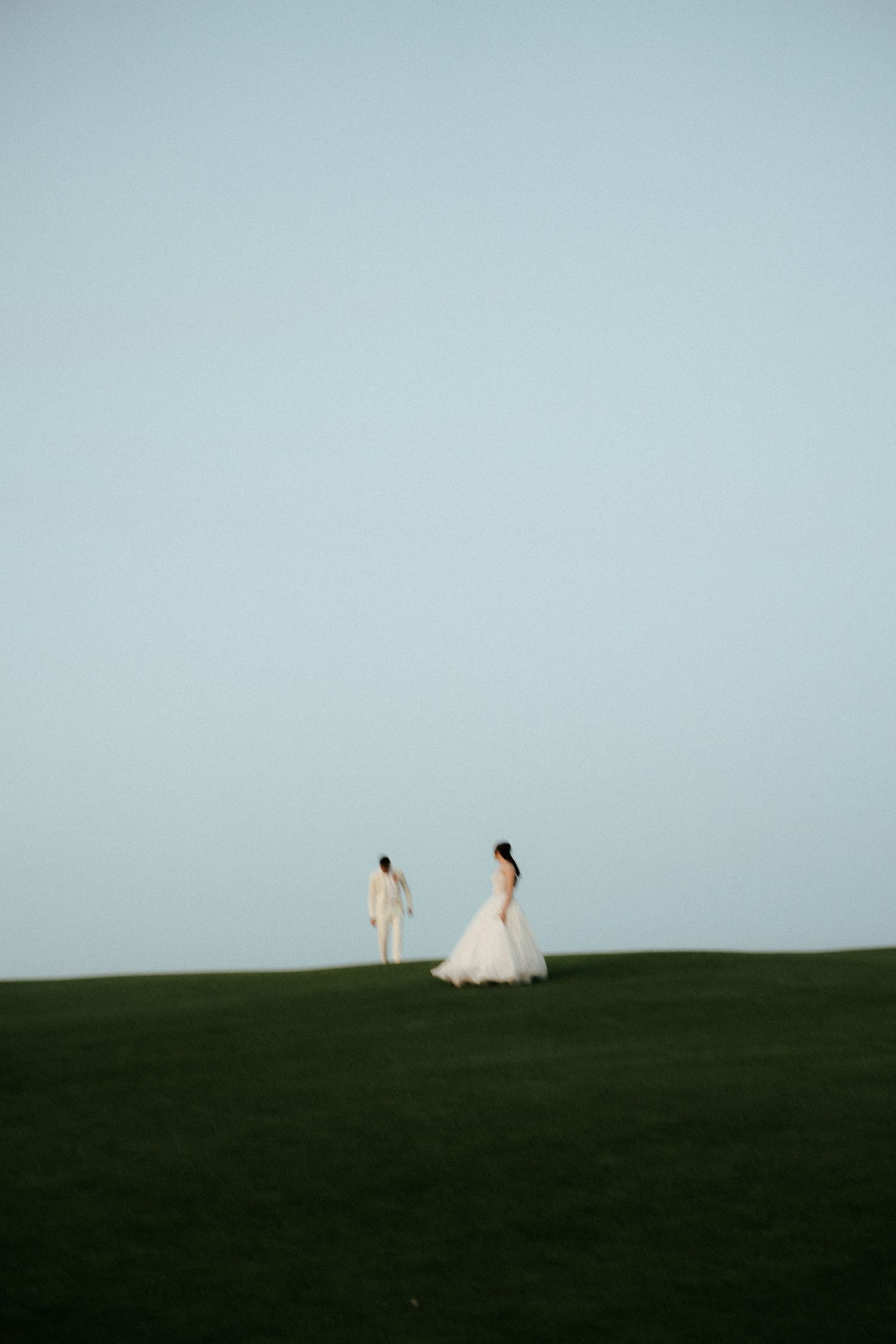 Two figures stand together on a grassy hilltop against a soft blue sky at dusk.