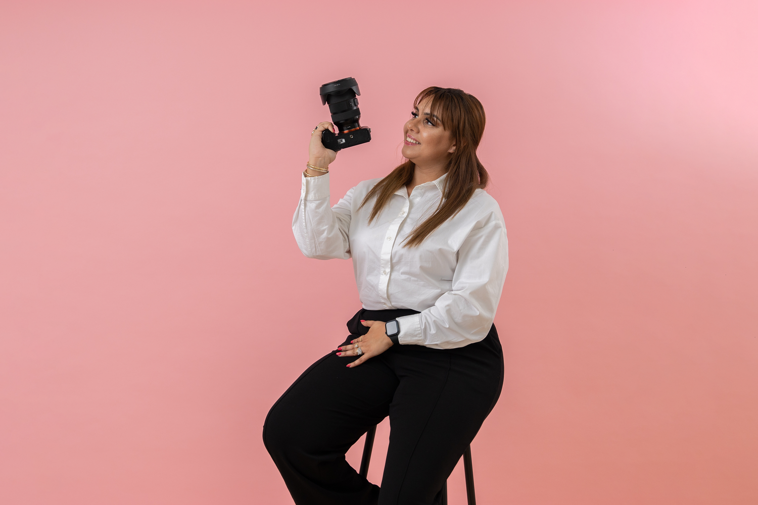 A woman in a white blouse and black pants sits on a stool holding a camera against a pink background.