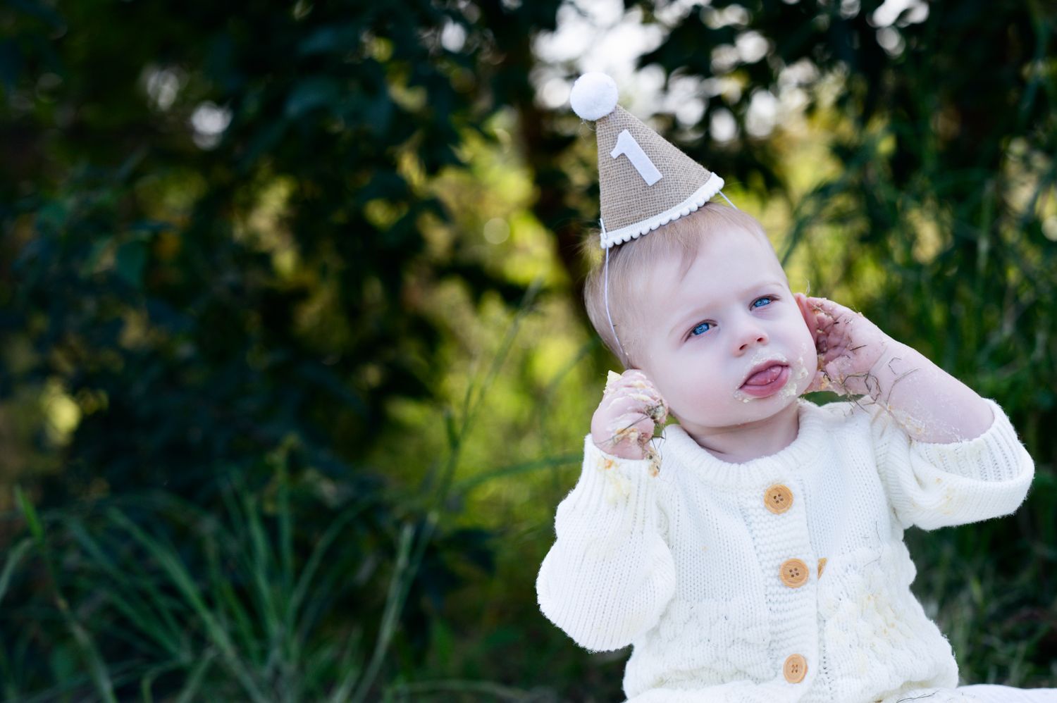 A baby wearing a white knit cardigan and party hat sits outdoors against a green natural background.