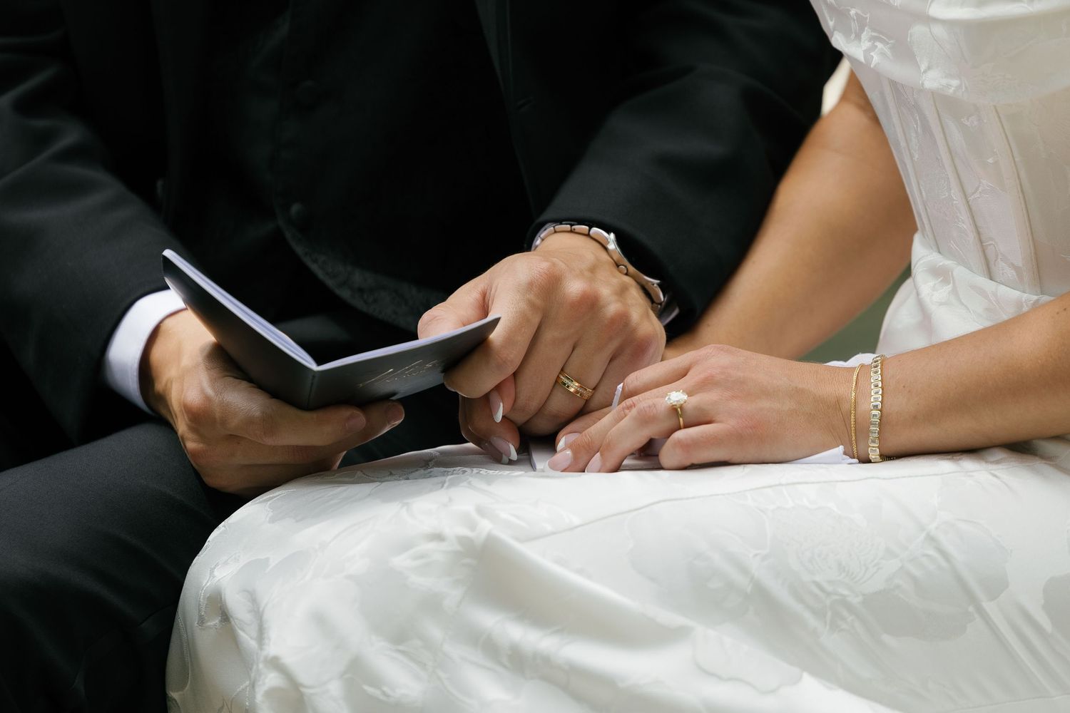 Close-up view of hands holding a ring during wedding ceremony, with bride in white dress and groom in black suit.