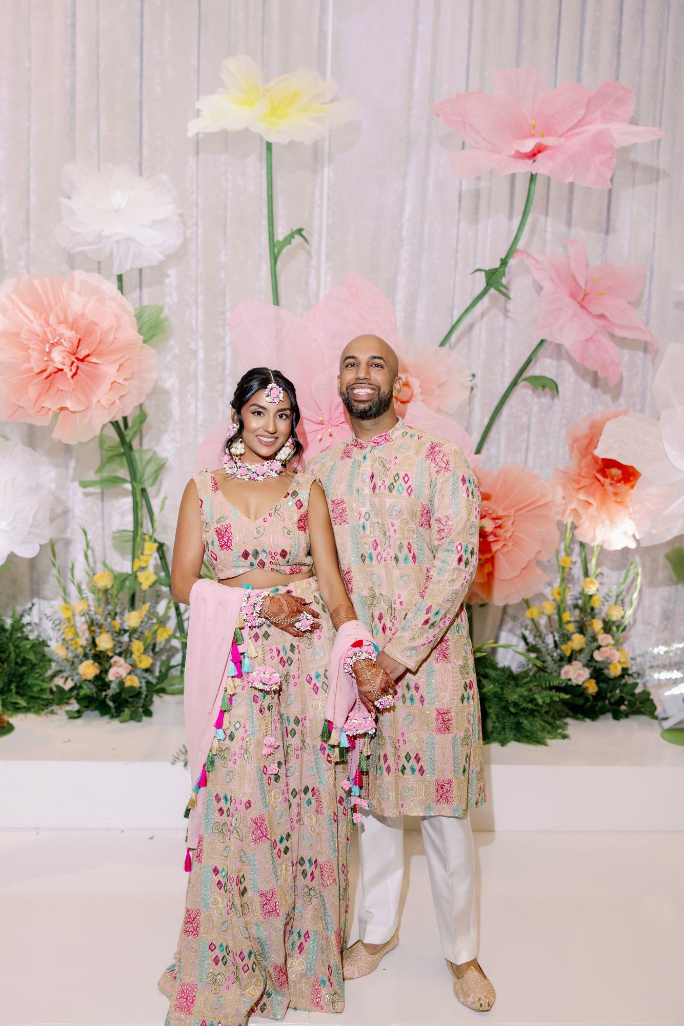 Couple in matching floral dresses pose against pink and white paper flower backdrop in dreamy spring photoshoot.