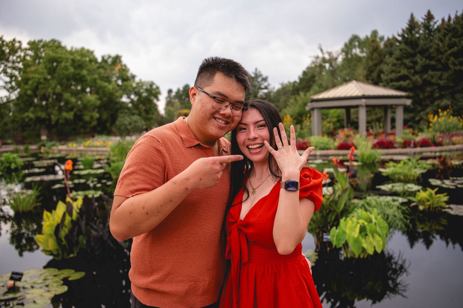 Couple stands by lakeside at sunset in park with gazebo, laughing and embracing during engagement photoshoot.