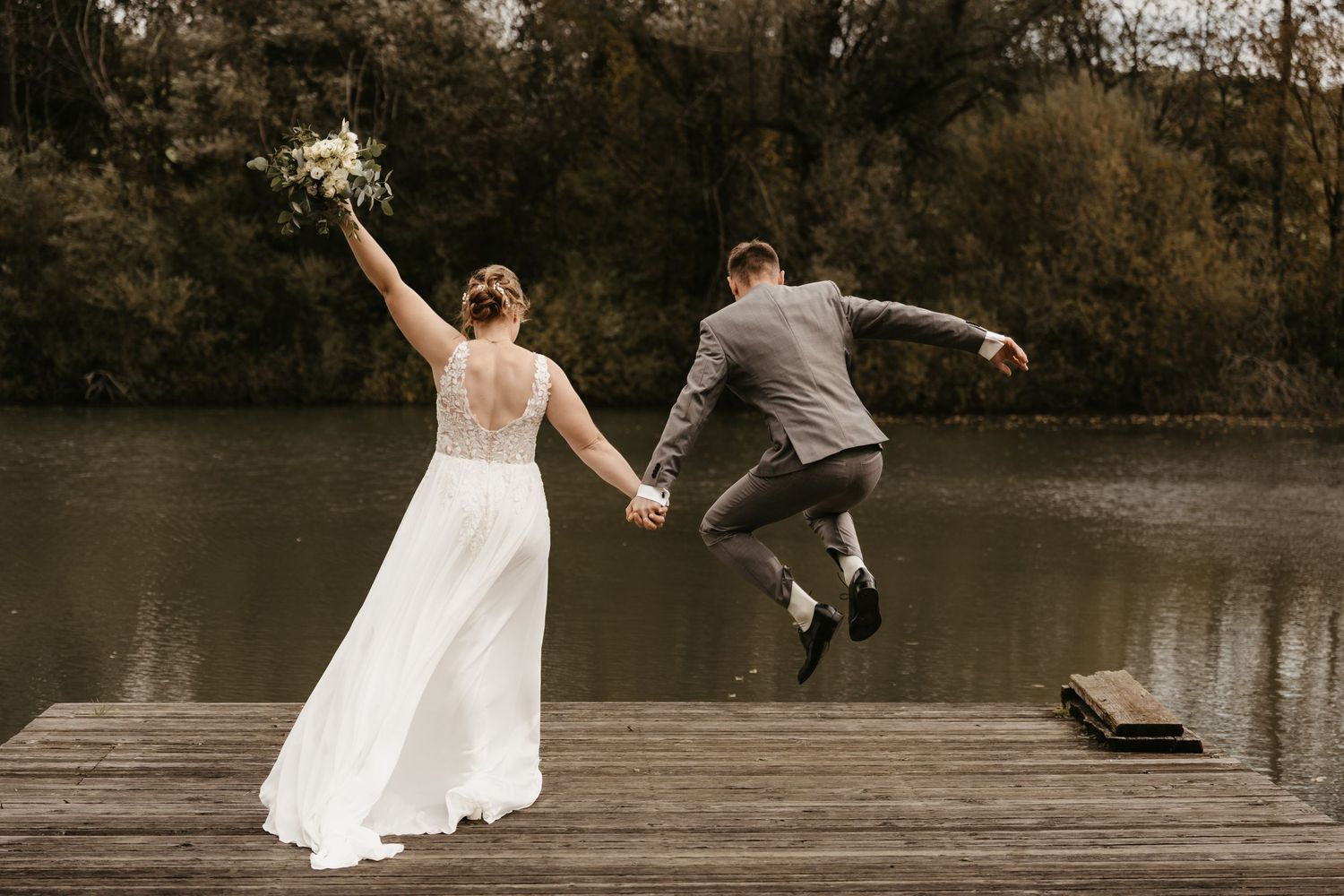 A couple in wedding attire dances and twirls together on a wooden dock by a serene lake at sunset.