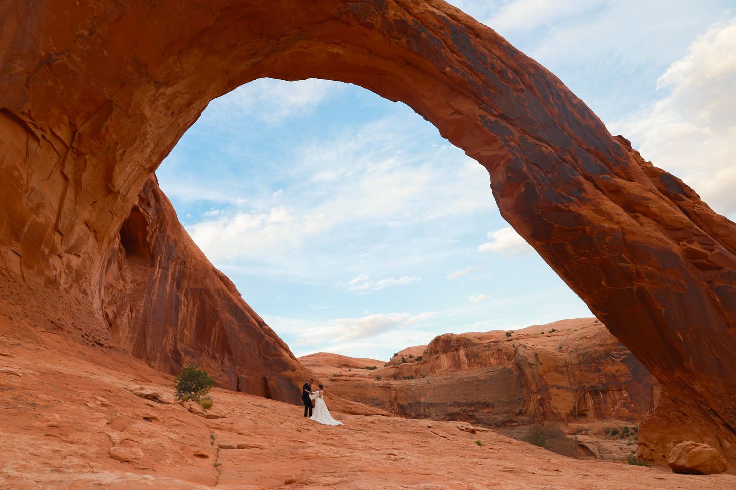A couple shares an intimate moment under a natural stone arch at night, illuminated by lantern light in the desert.