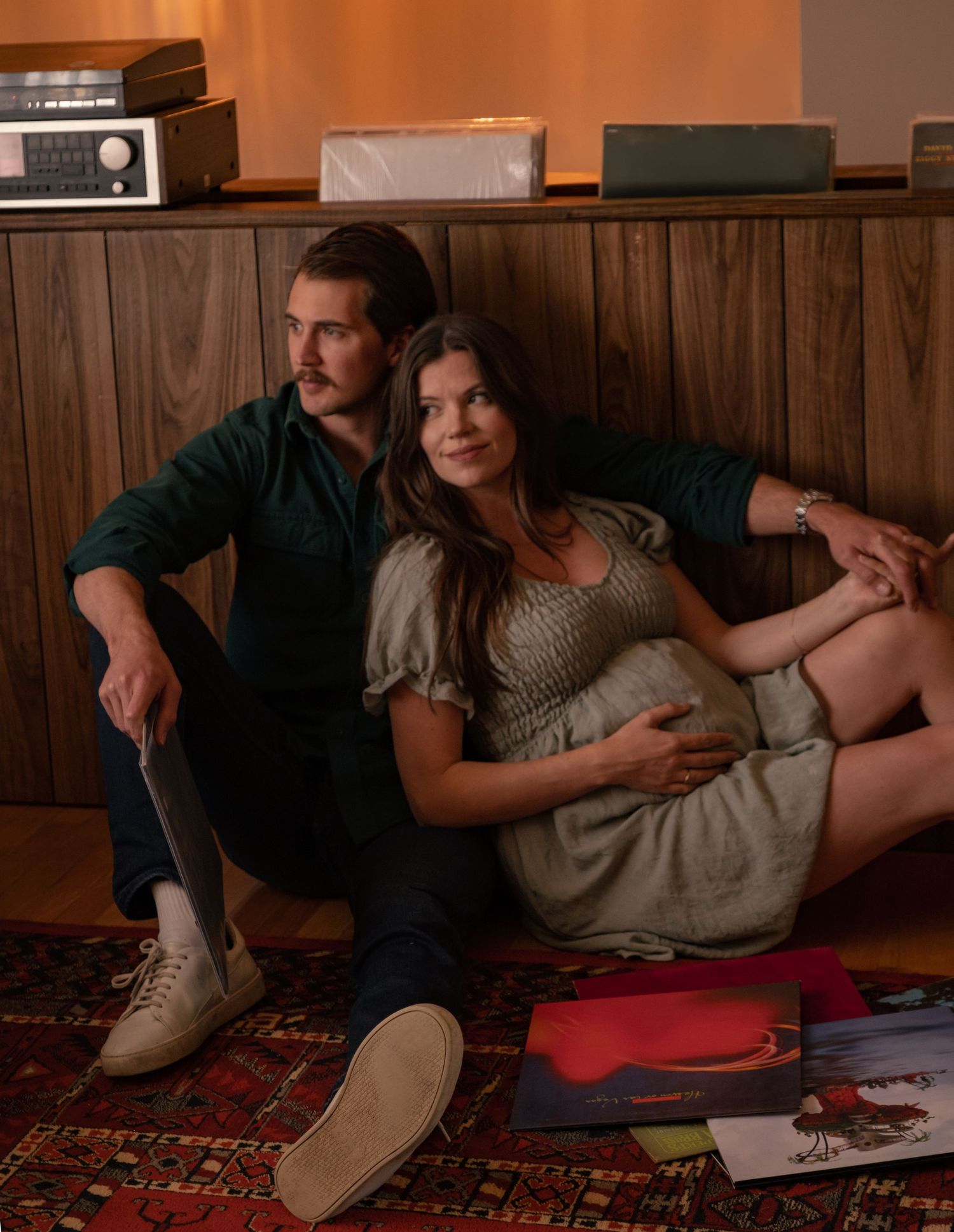 Two people relax together on a patterned rug in a cozy room with wood paneled walls.