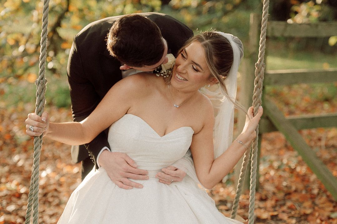 A couple in wedding attire sits together on a hilltop overlooking a scenic valley landscape at sunset.