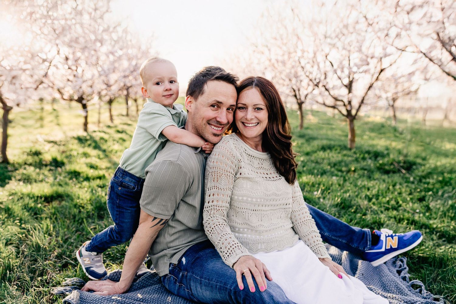 Mama, Papa und ihr kleiner Sohn sitzen auf der Wiese zwischen blühenden Marillenbäumen und lachen in die Kamera.