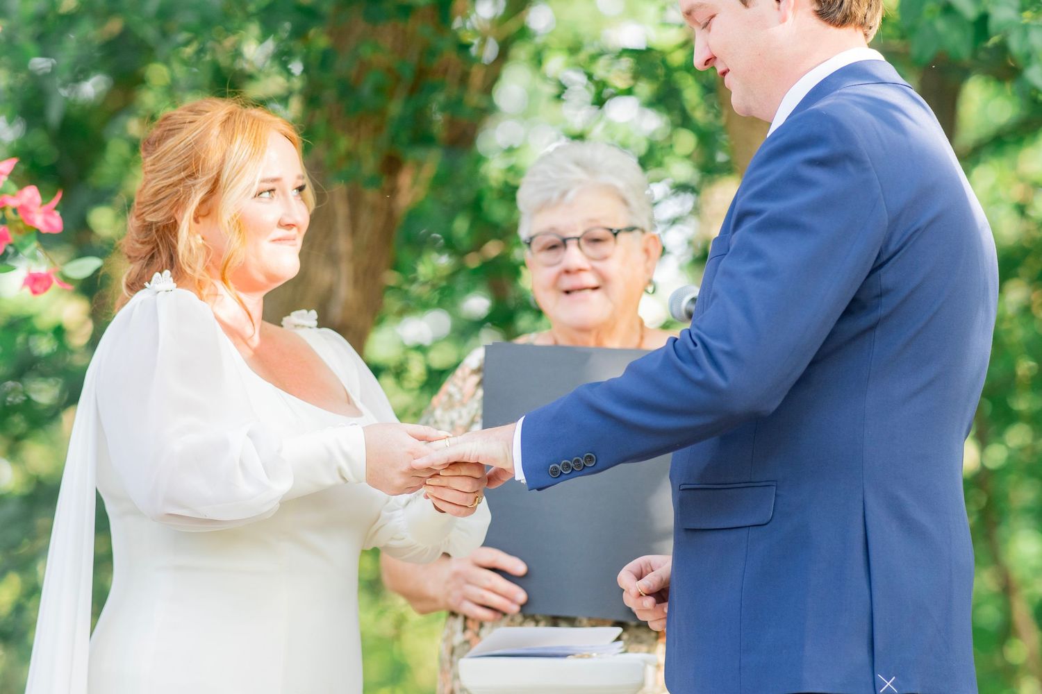 Bride and groom exchange rings during an outdoor wedding ceremony with an officiant present.
