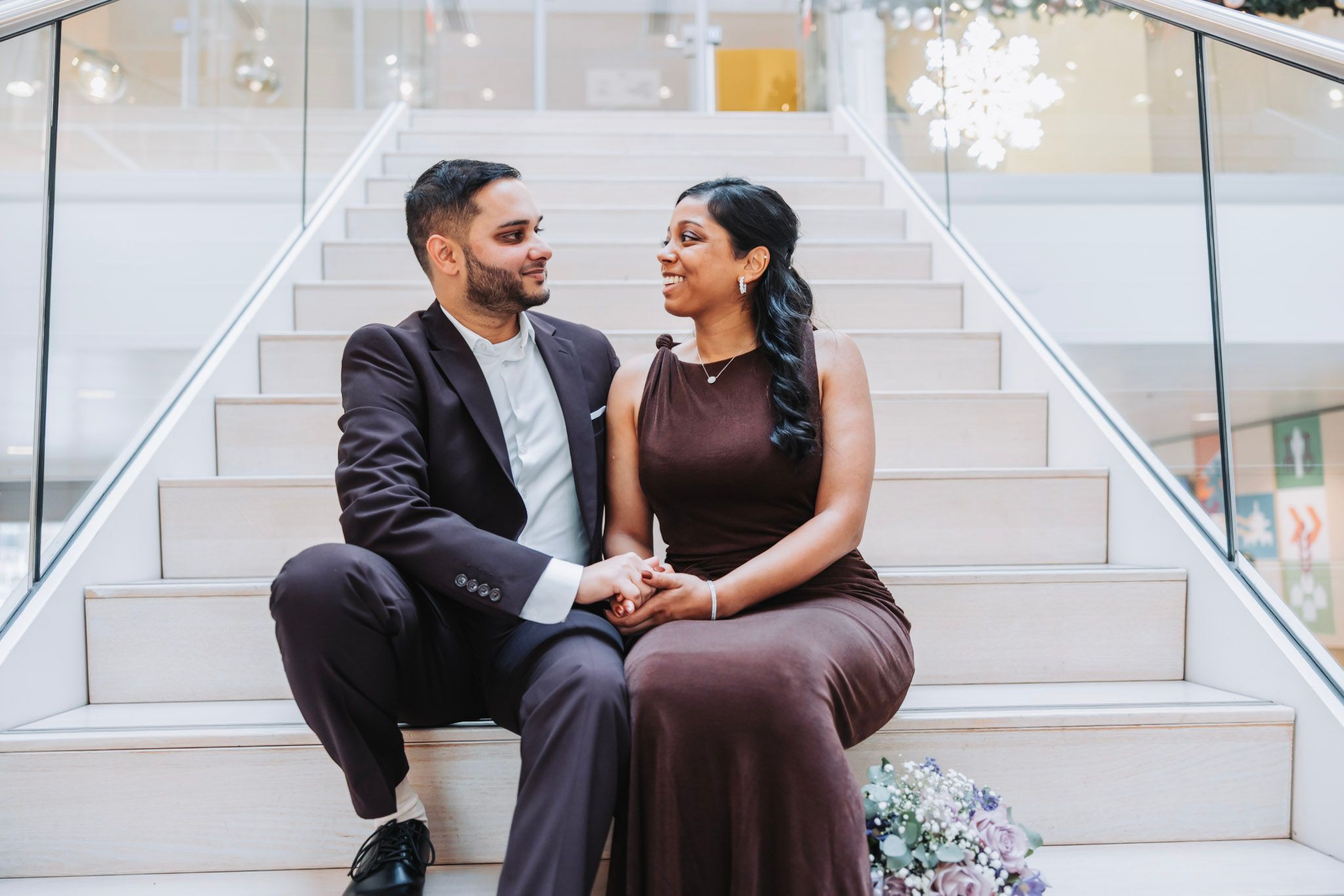 A couple in formal attire sits and chats on bright staircase inside Gemeente Hoofddorp looking at each other.