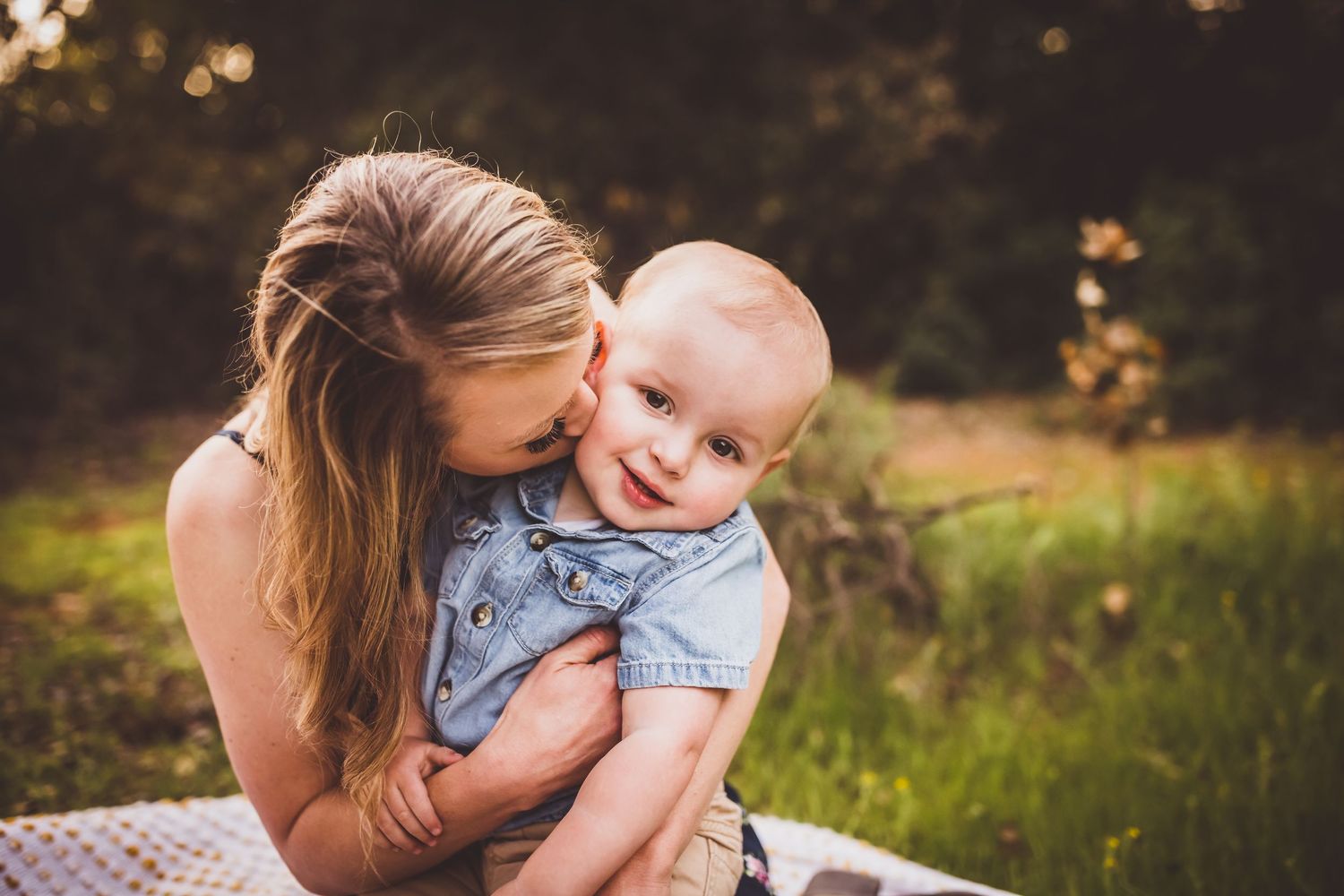 A joyful moment captured outdoors as a baby in denim laughs while being held in a grassy setting at sunset.