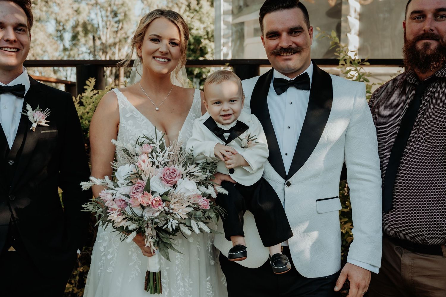 Wedding party in formal attire poses outdoors with elegant pink and white floral bouquets.