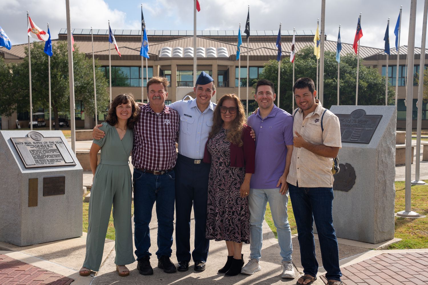 A group of people standing together outdoors in front of a memorial or monument with flags waving in the background.