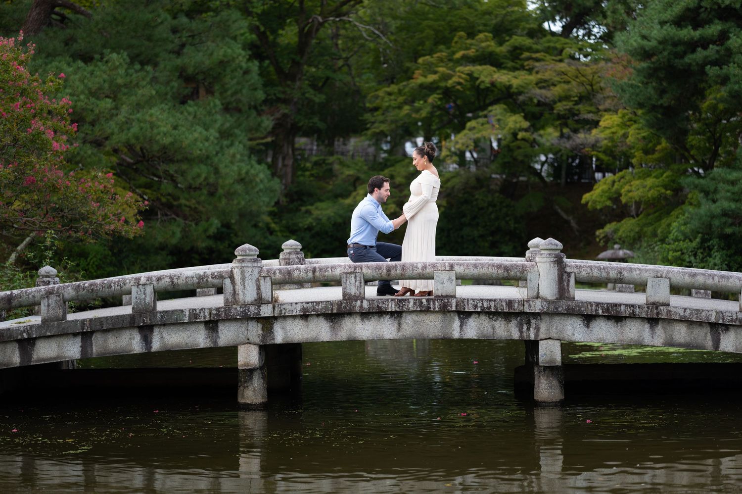 Man proposing on a stone bridge over a tranquil pond in a lush Japanese garden.