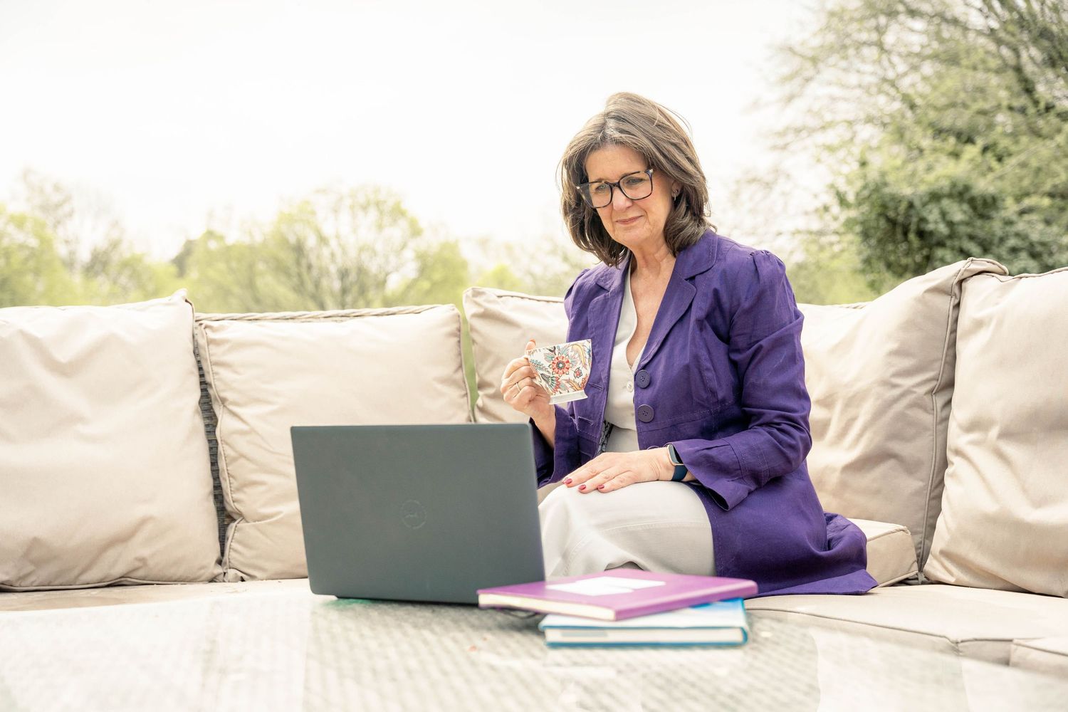 personal branding shoot Brighton. a woman sits outside on garden furniture with an open laptop and note books on the table she holds a cup