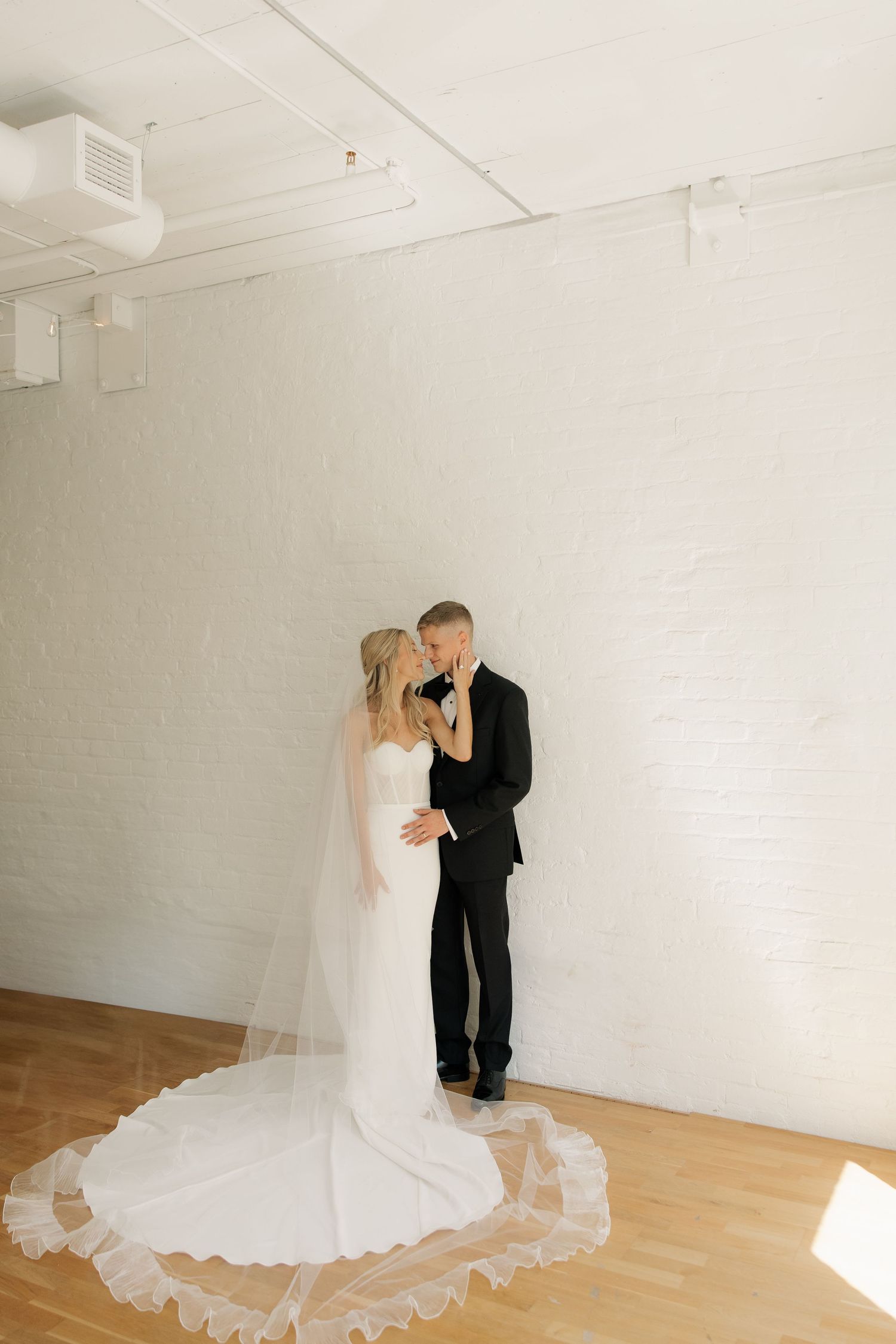 A couple in formal wedding attire share an intimate moment in a minimalist white studio space with wooden floors.
