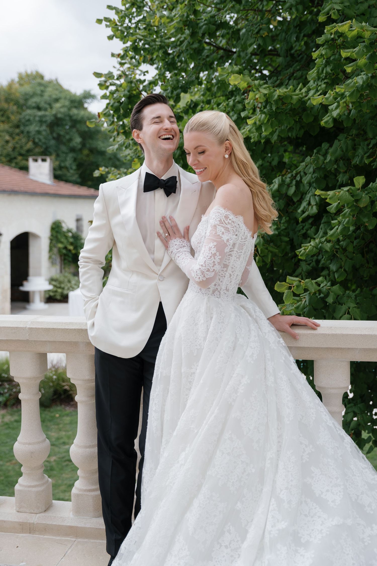 A romantic wedding couple in white attire share tender moments on a terrace with stone balustrades and lush greenery.