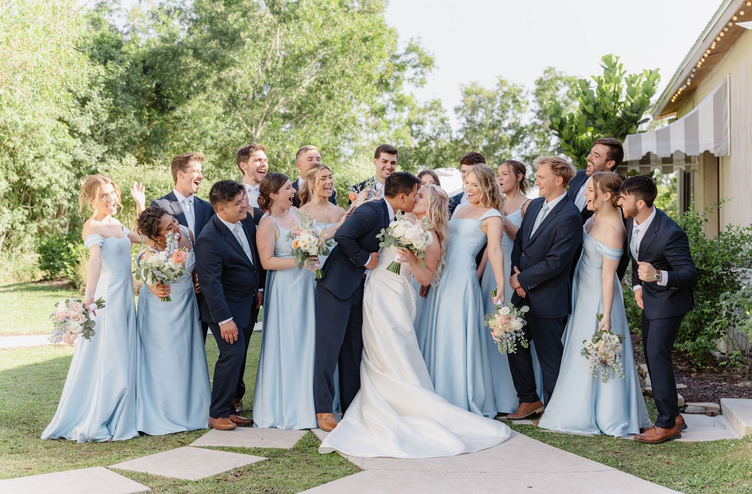 Outdoor wedding ceremony guests gather on white chairs in front of a rustic barn on a sunny afternoon.