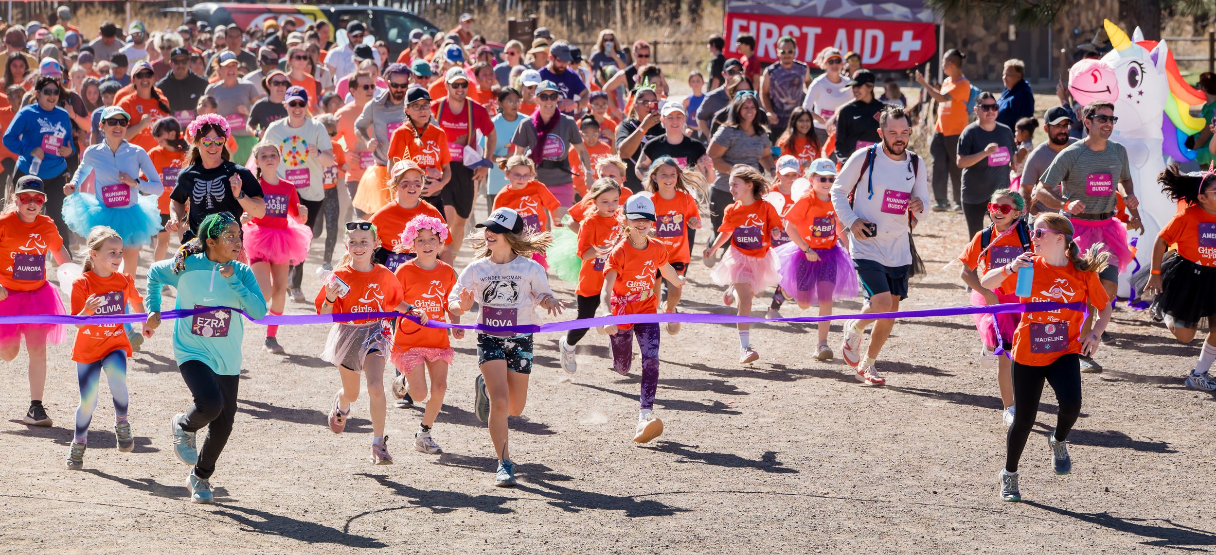 A large crowd of runners wearing orange shirts participate in a community race event on a sunny day.