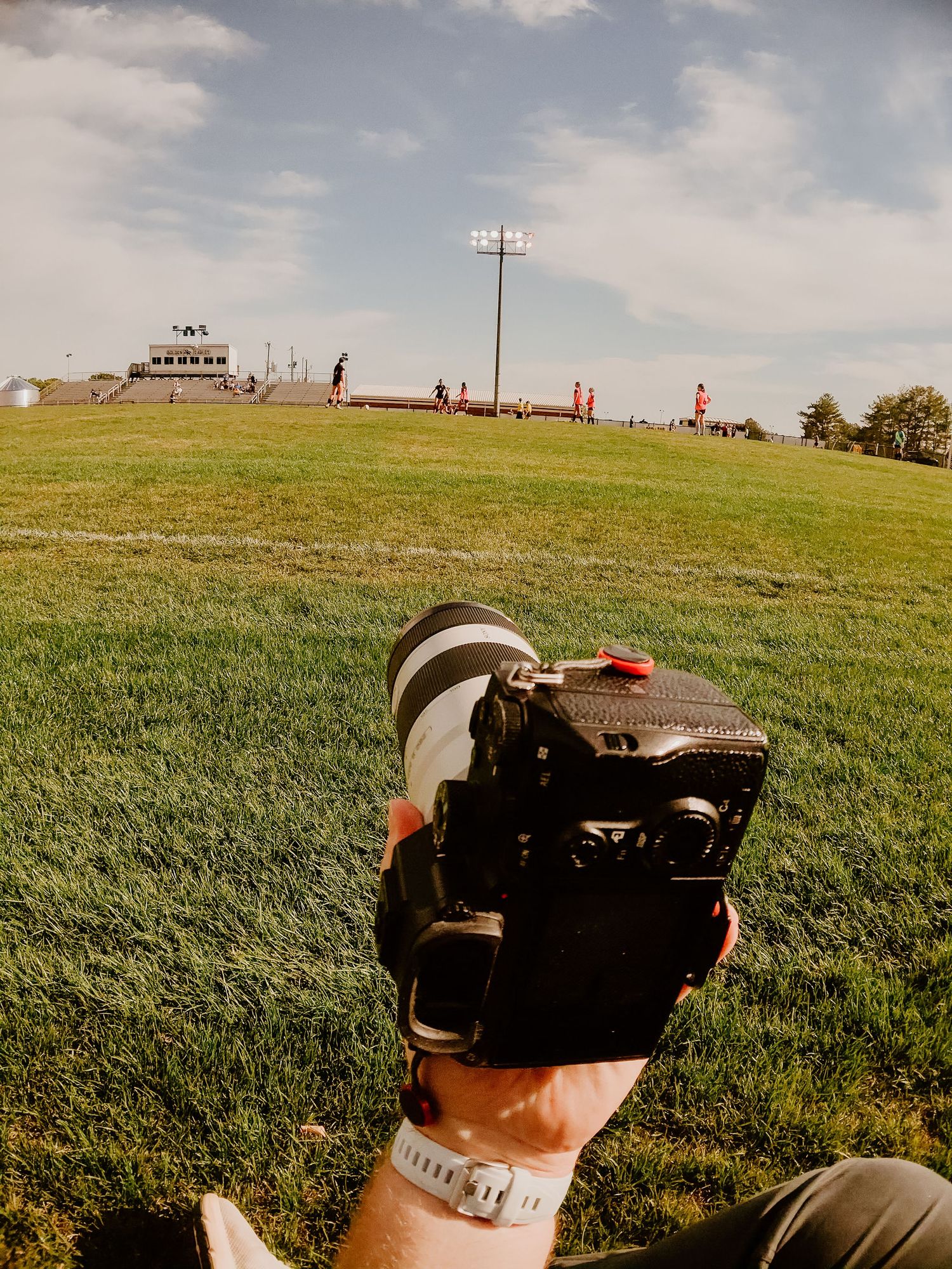 Photographer holding a Canon DSLR camera with telephoto lens on a green sports field under a blue sky.