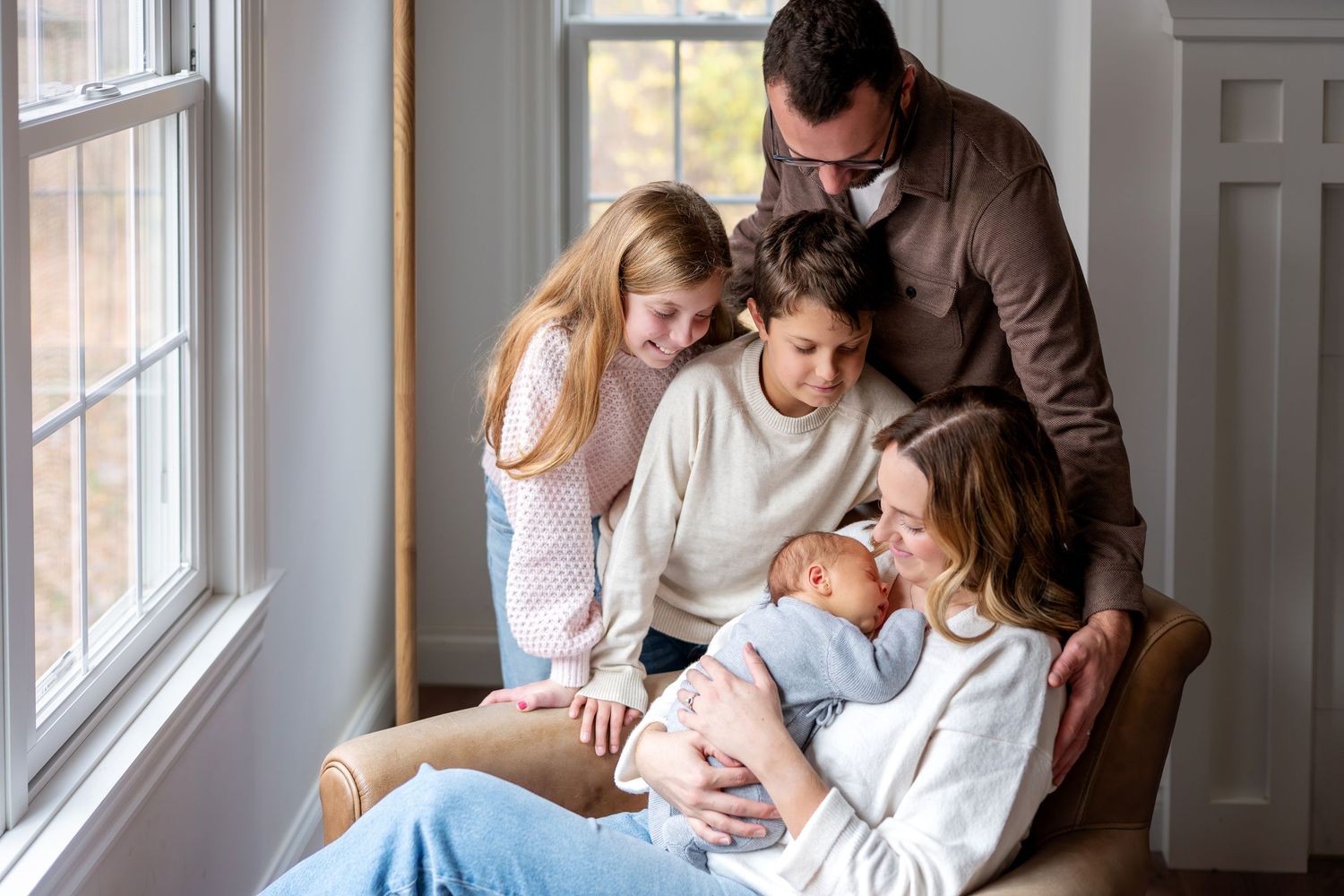 Family of five gathered around mother holding newborn baby by a bright window in a cozy home.