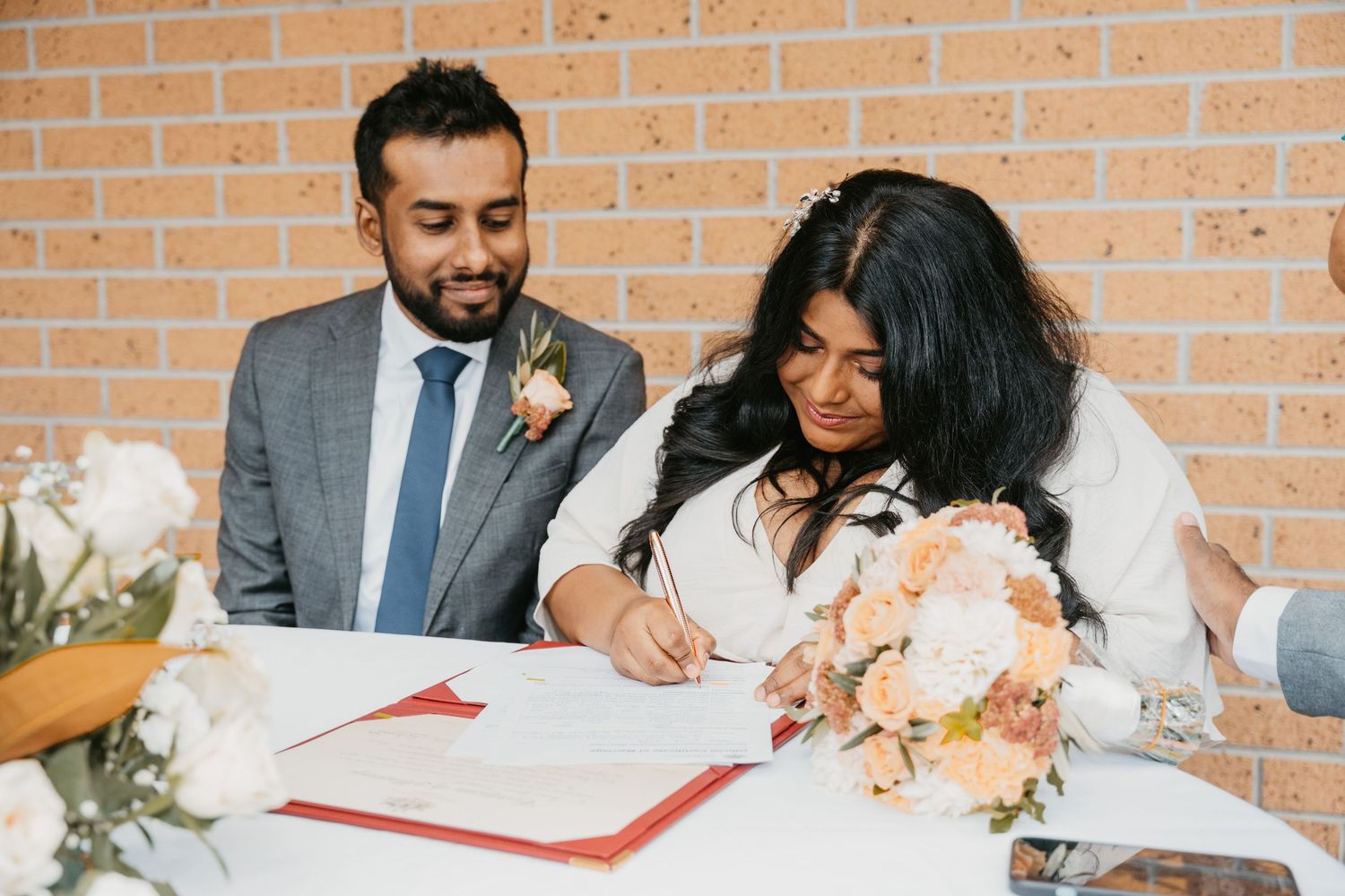 A couple signs marriage documents at a ceremony with peach and white floral decorations on a brick wall background.