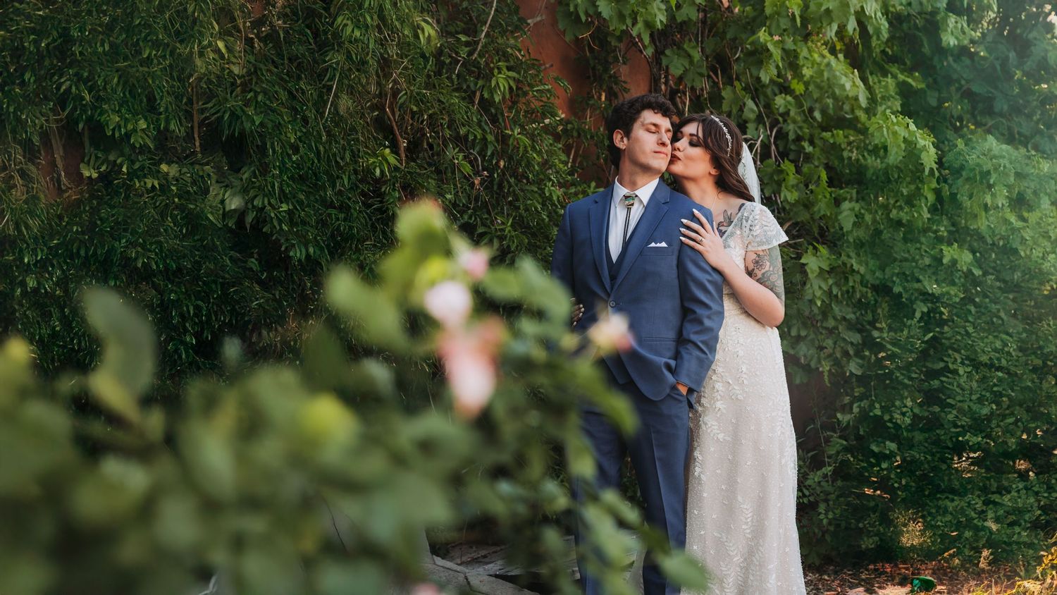 A couple poses together in an outdoor garden setting with lush greenery, sunshine, and elegant formal attire.