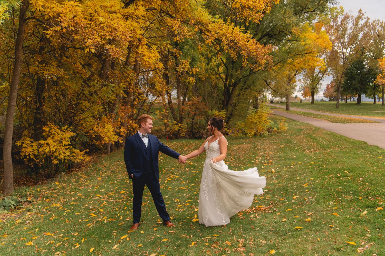 A couple walks hand in hand through a scenic autumn landscape with golden trees and fallen leaves along a path.