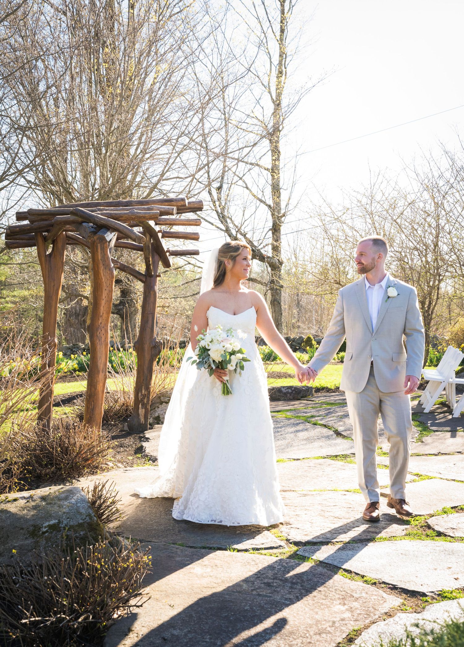 Bride and groom holding hands in a garden wedding venue with rustic wooden arbor and stone pathway on a sunny day.