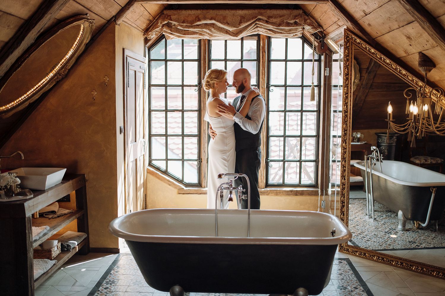 Romantic couple posing by vintage bathtub near large window in rustic attic setting.