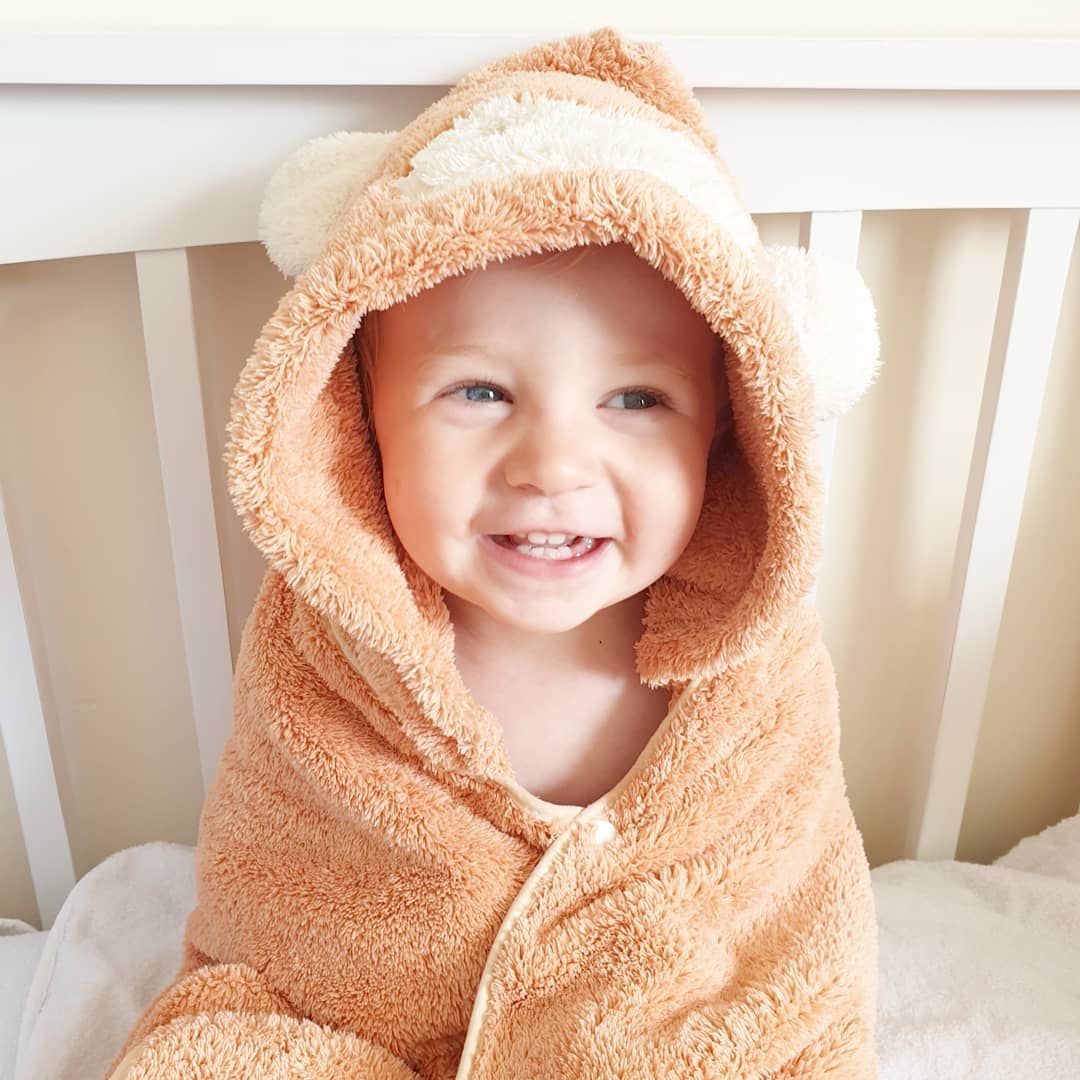 Smiling toddler wrapped in peach hooded towel sitting in white crib.