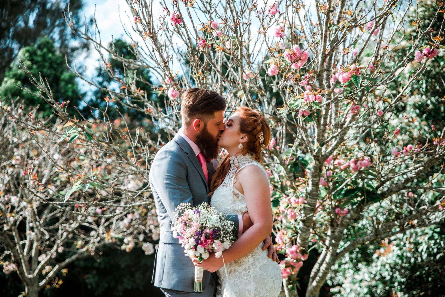 A couple shares an intimate moment among blooming spring trees with a floral bouquet.