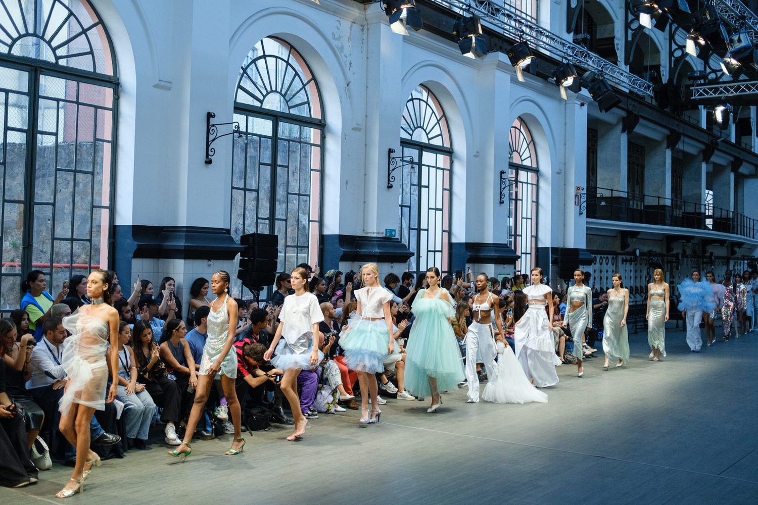 Fashion show models walk a runway in an industrial space with dramatic architectural details and natural lighting.