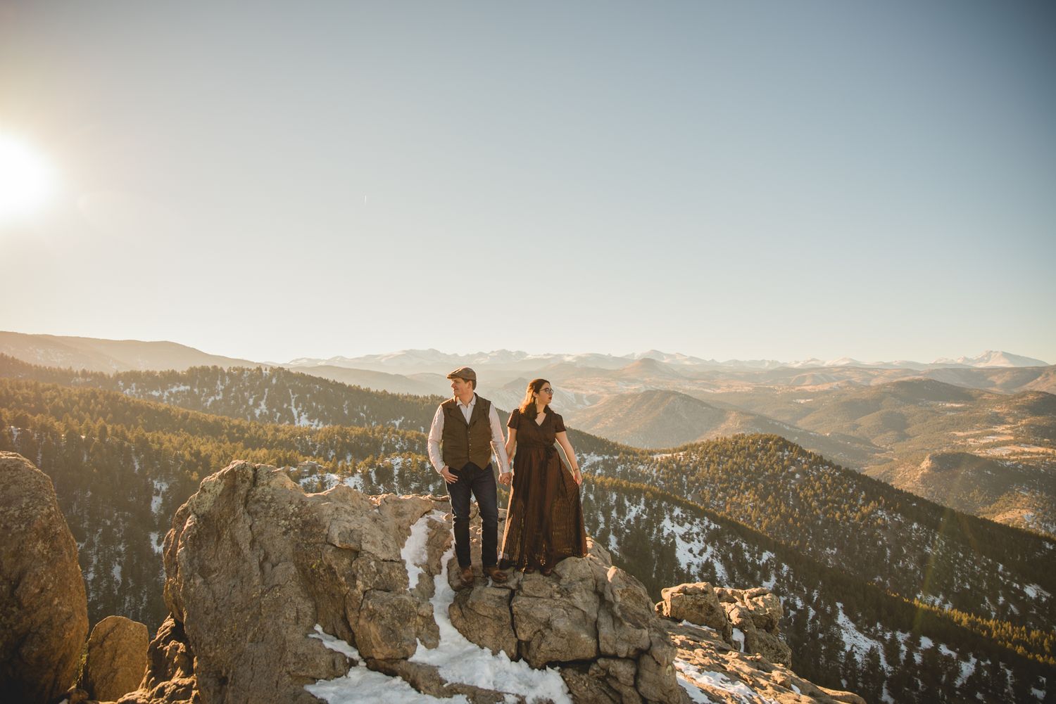 A couple stands on a rocky mountain overlook with snow-capped peaks and valleys in the background.