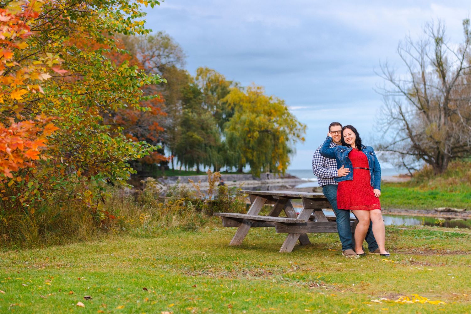 Autumn engagement photo at a lakeside park with colorful fall foliage.