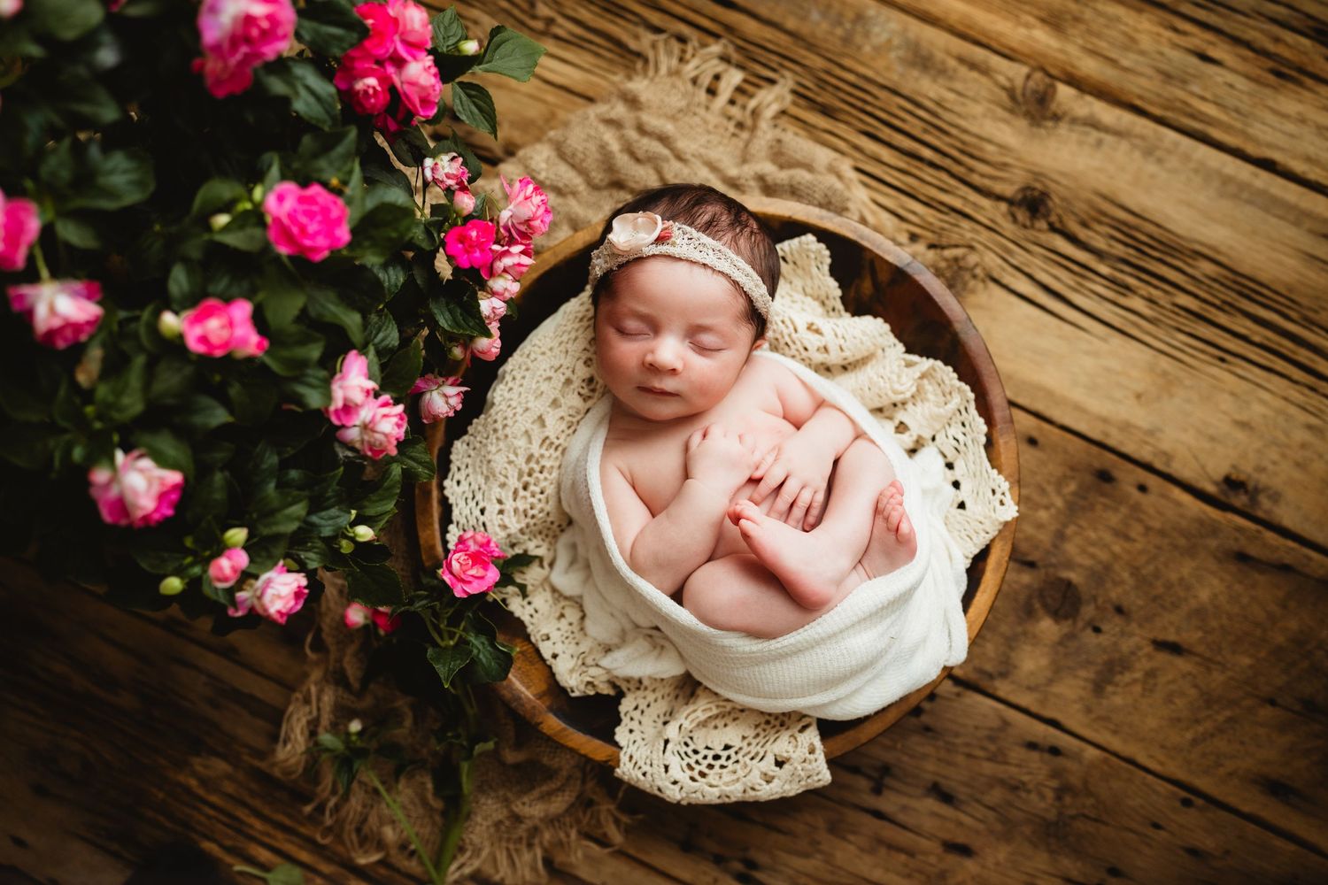 A sleeping newborn in a white wrap with pink flowers and wooden backdrop.