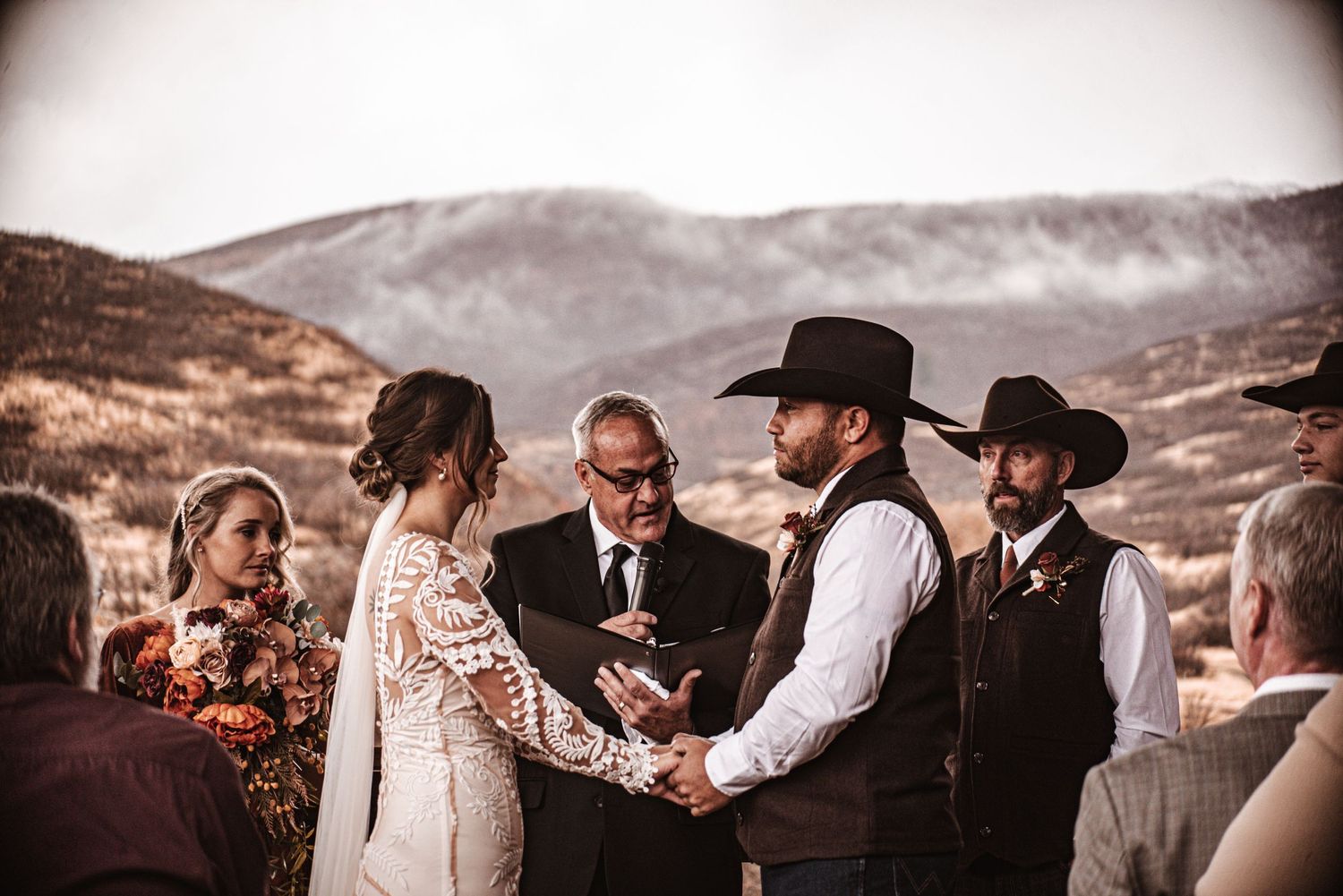 A western-style outdoor wedding ceremony takes place against a backdrop of mountain scenery.