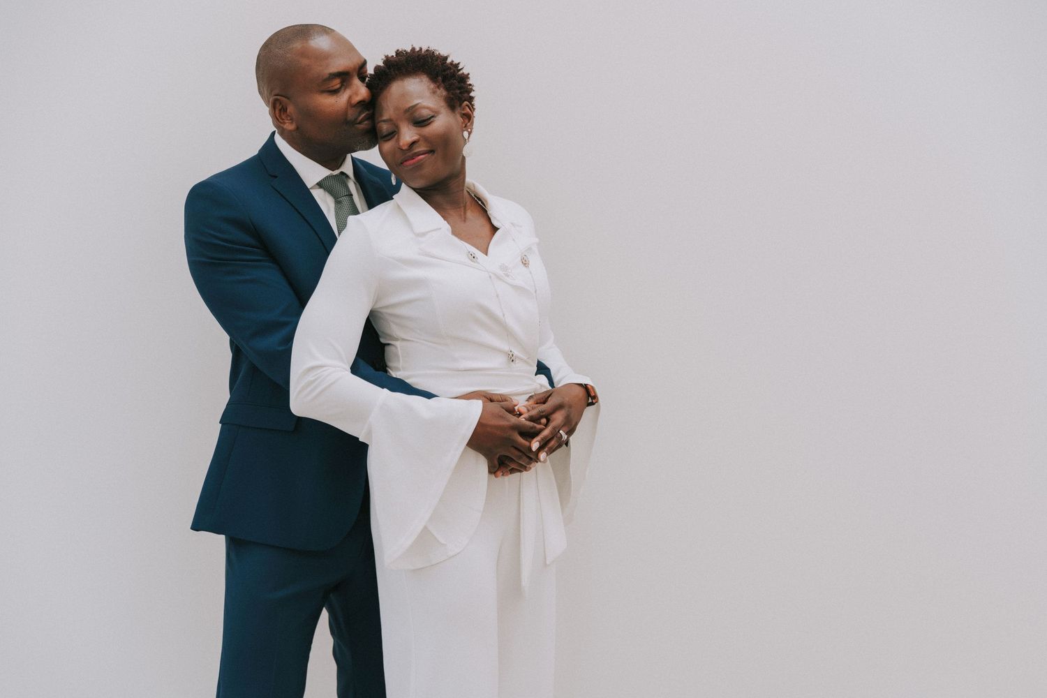 A romantic couple embraces against a gray background, wearing elegant white and navy formal attire.