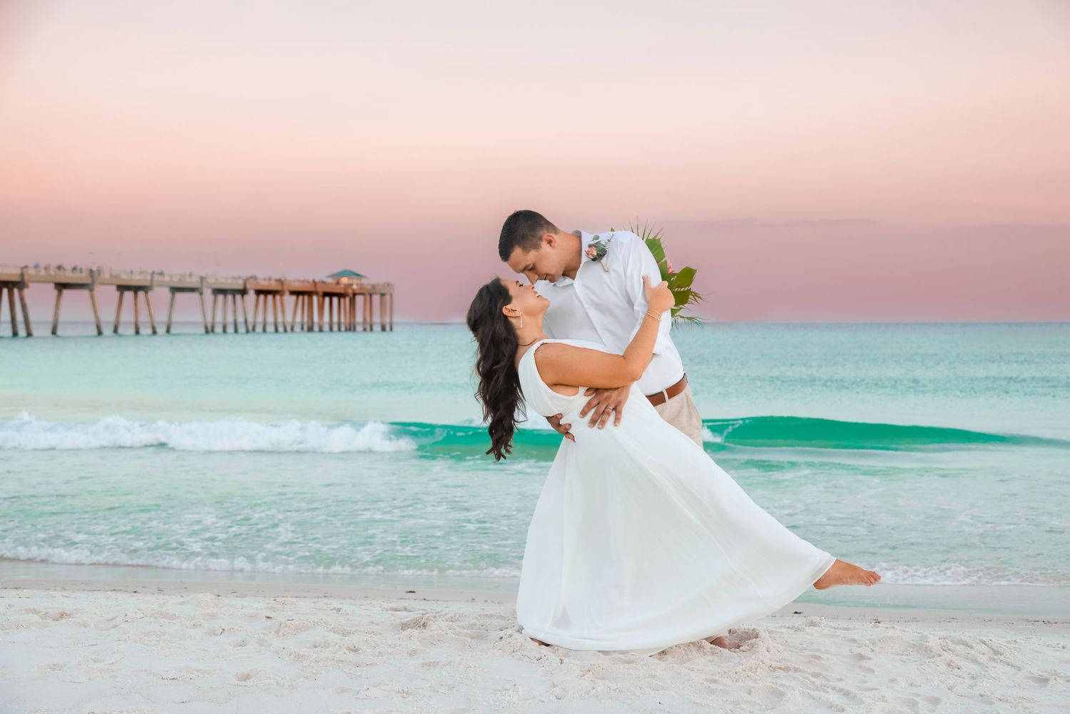 A romantic dance dip on a white sandy beach with a pier and turquoise waters in the background.
